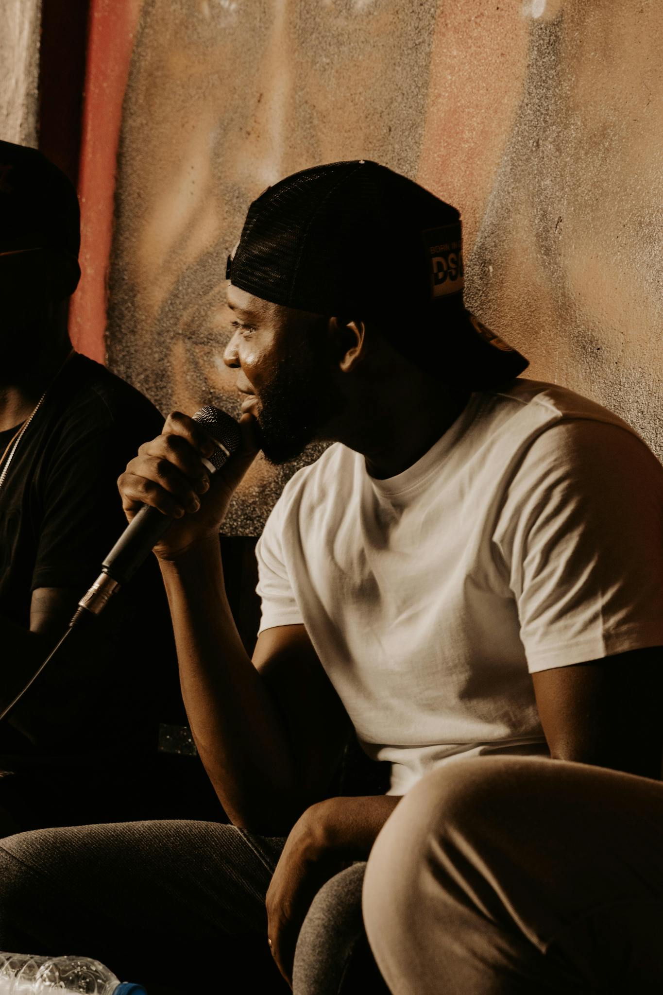 Black man in cap holding microphone in casual setting, singing or rapping.